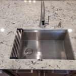 Stainless steel kitchen sink with a tall curved faucet and soap dispenser on a white granite countertop.