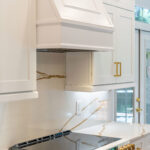 White kitchen featuring a built-in hood and white cabinets with gold handles.